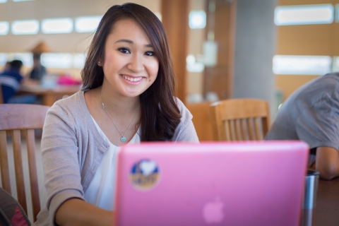 Student sitting with pink laptop in Kolligian Library and smiling at camera