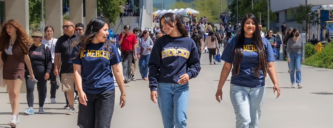 Three students wearing UC Merced shirts in the center of the frame walking in the middle of the busy UC Merced campus